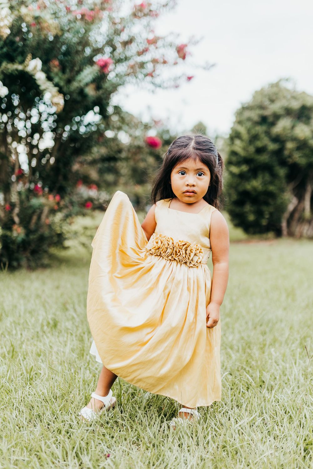 A young child poses in a cream-colored party dress with gold embellishments in a lush garden setting.
