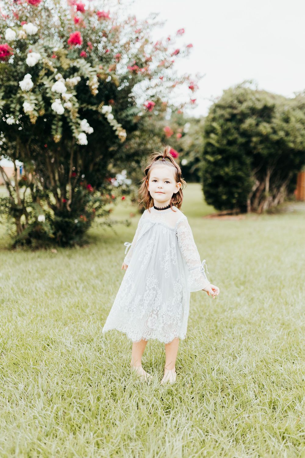 A small child in a white lace dress twirls in a grassy garden with blooming rose bushes in the background.