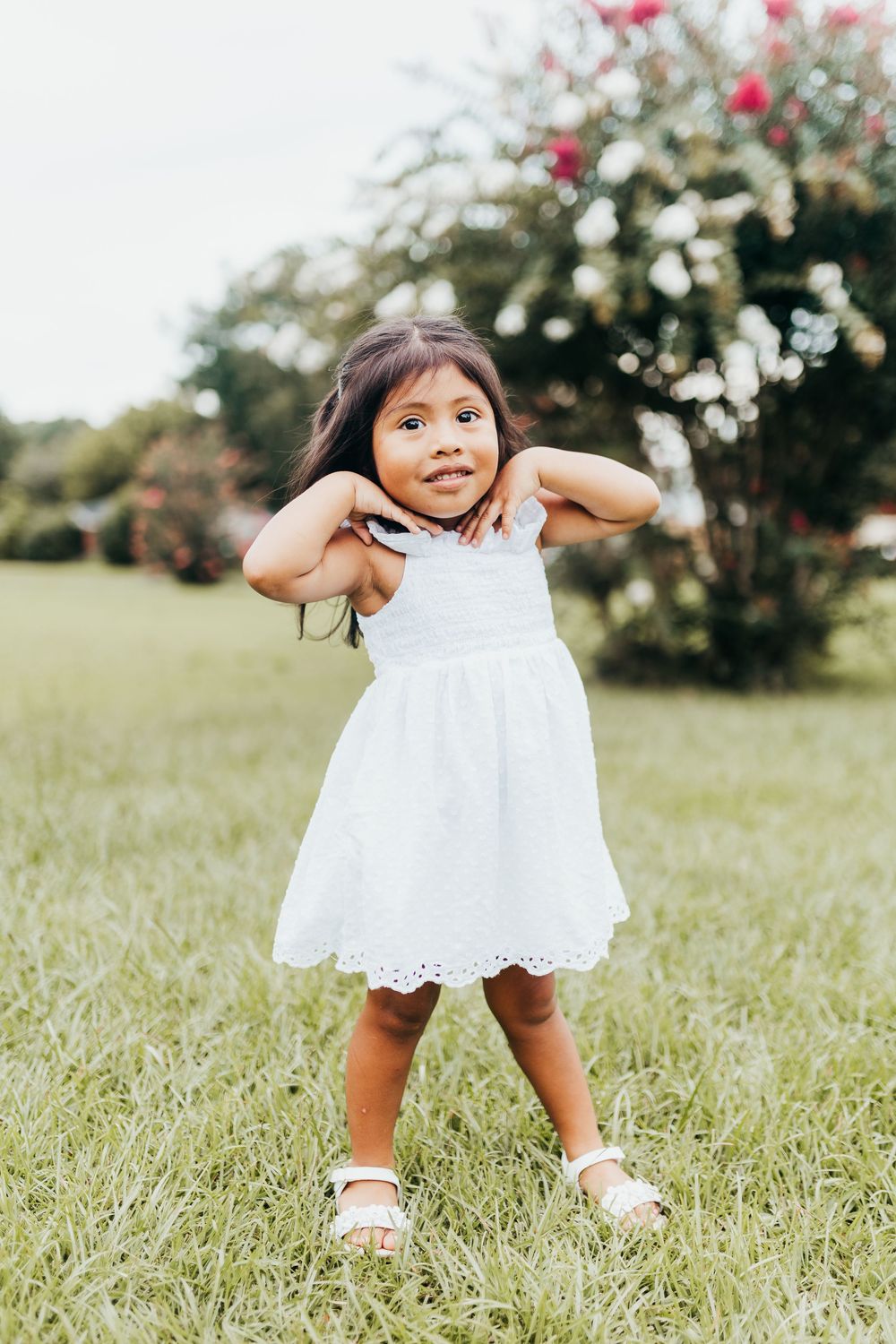 A playful candid moment in a white summer dress against a sunny garden backdrop with blooming flowers.