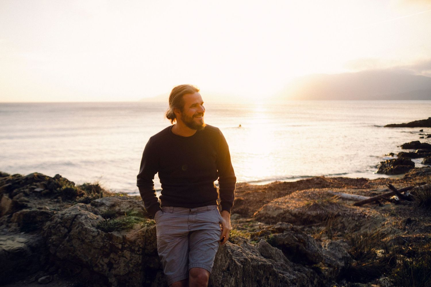 Person standing on rocky coastline at sunset with ocean view and warm golden light in background.