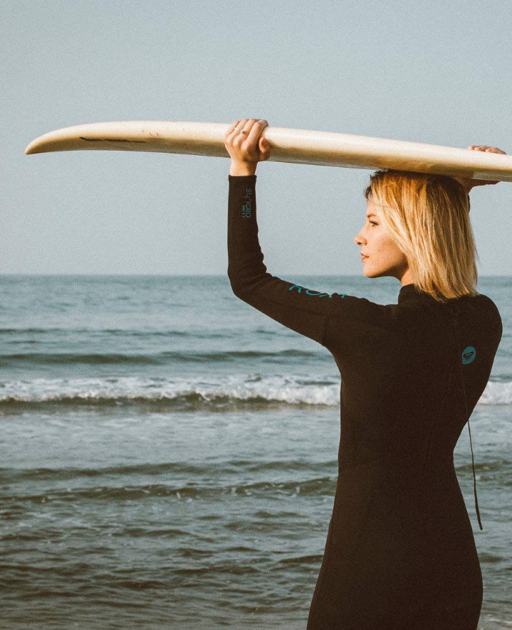 A surfer in a black wetsuit holds a surfboard overhead while standing on the beach by the ocean.