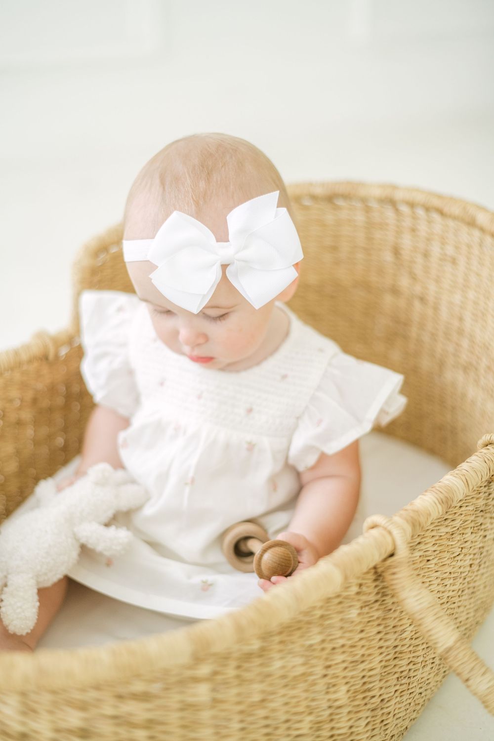 Baby with white bow headband sitting in wicker bassinet with toys.
