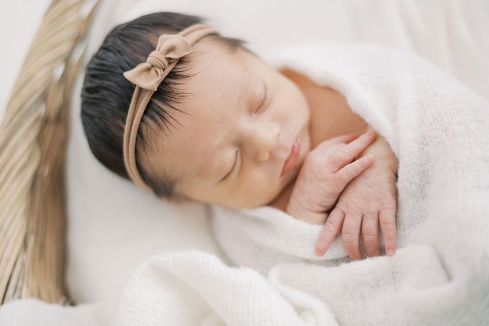 Close-up of a sleeping newborn baby girl with dark hair wearing a tan bow headband, wrapped in white fabric.