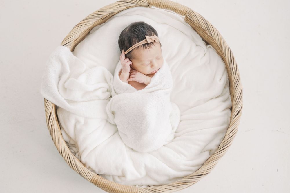 Sleeping newborn baby girl swaddled in white wrap rests peacefully in a round wicker basket with soft bedding.