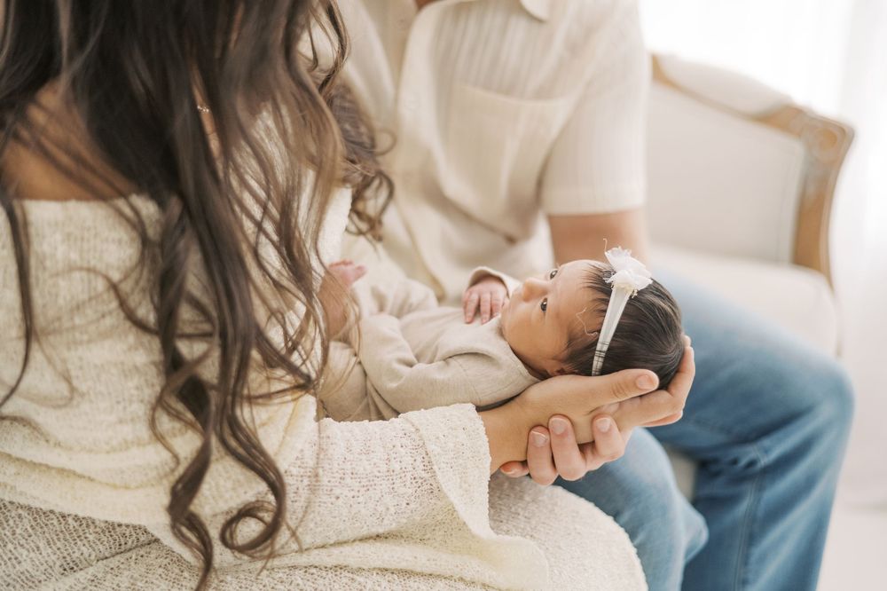 New parents gently cradle their sleeping newborn baby, mother in cream lace dress, father in beige shirt and jeans.