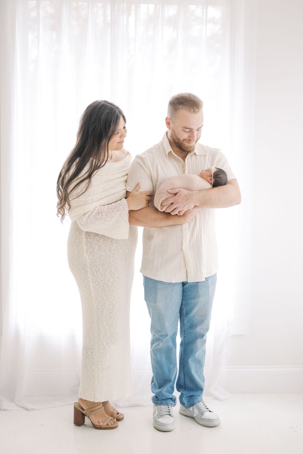 New parents stand by sheer white curtains, father cradling newborn as mother in white lace dress looks on lovingly.