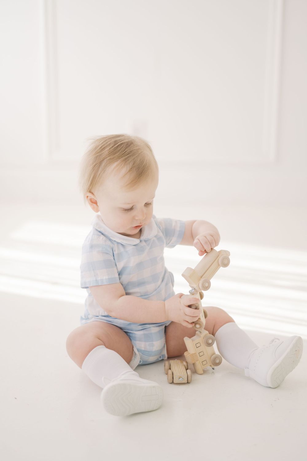 Adorable baby boy in blue gingham romper sits on white floor playing with a wooden toy train set.