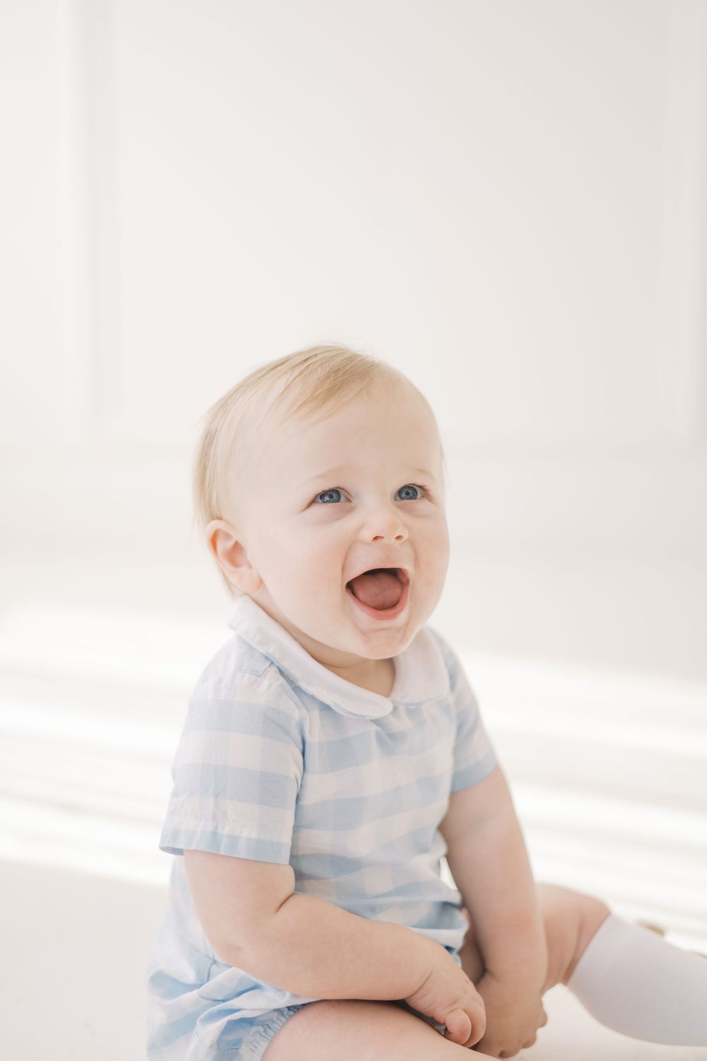 Laughing baby in blue gingham romper sitting on white floor.