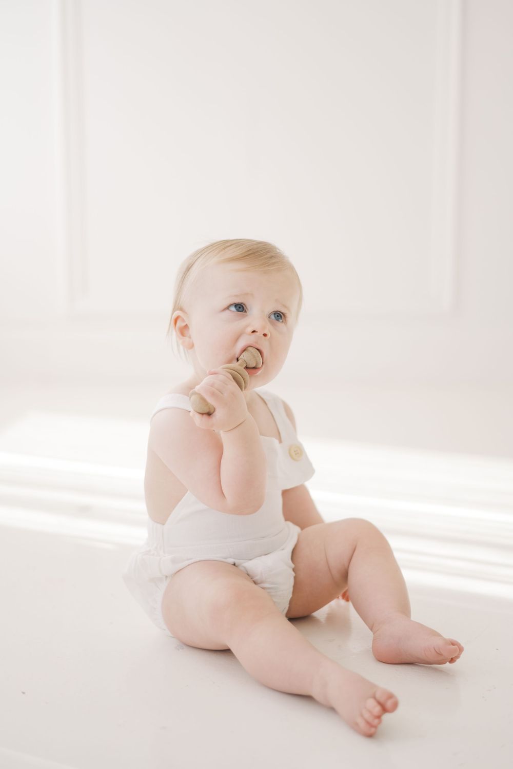 Cute baby in white romper sits on bright white floor, chewing on a wooden toy while looking upward curiously.