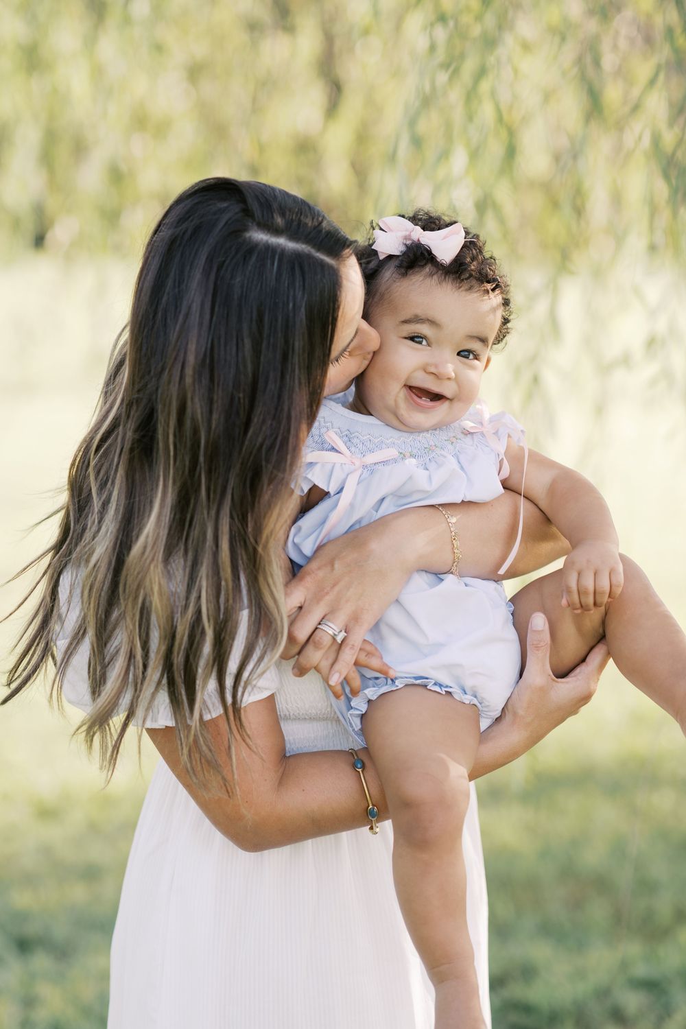 A mother kisses her laughing baby girl with a pink bow in a sun-drenched outdoor setting.