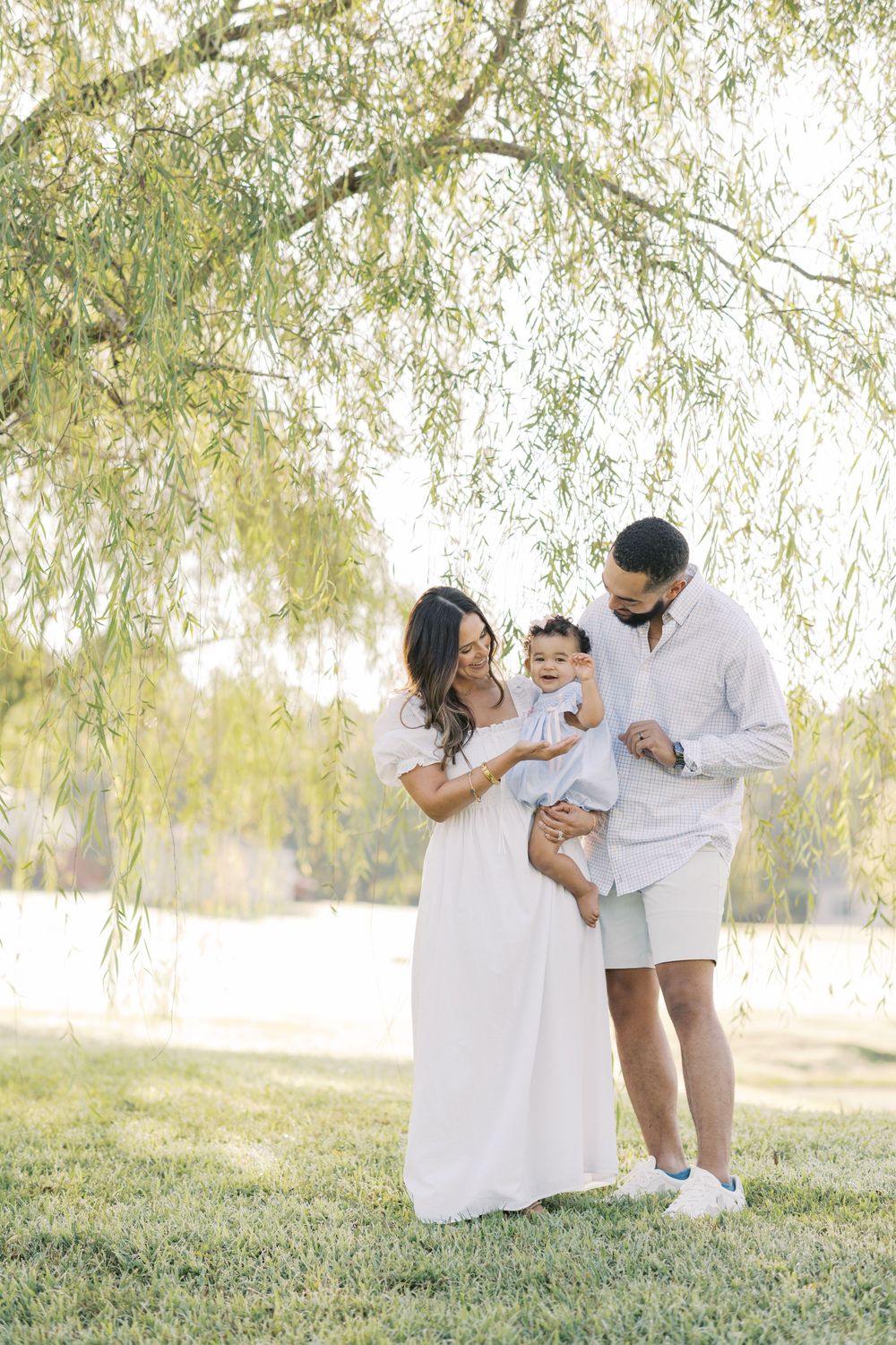 A family of three stands under a weeping willow tree, parents smiling at their baby outdoors.