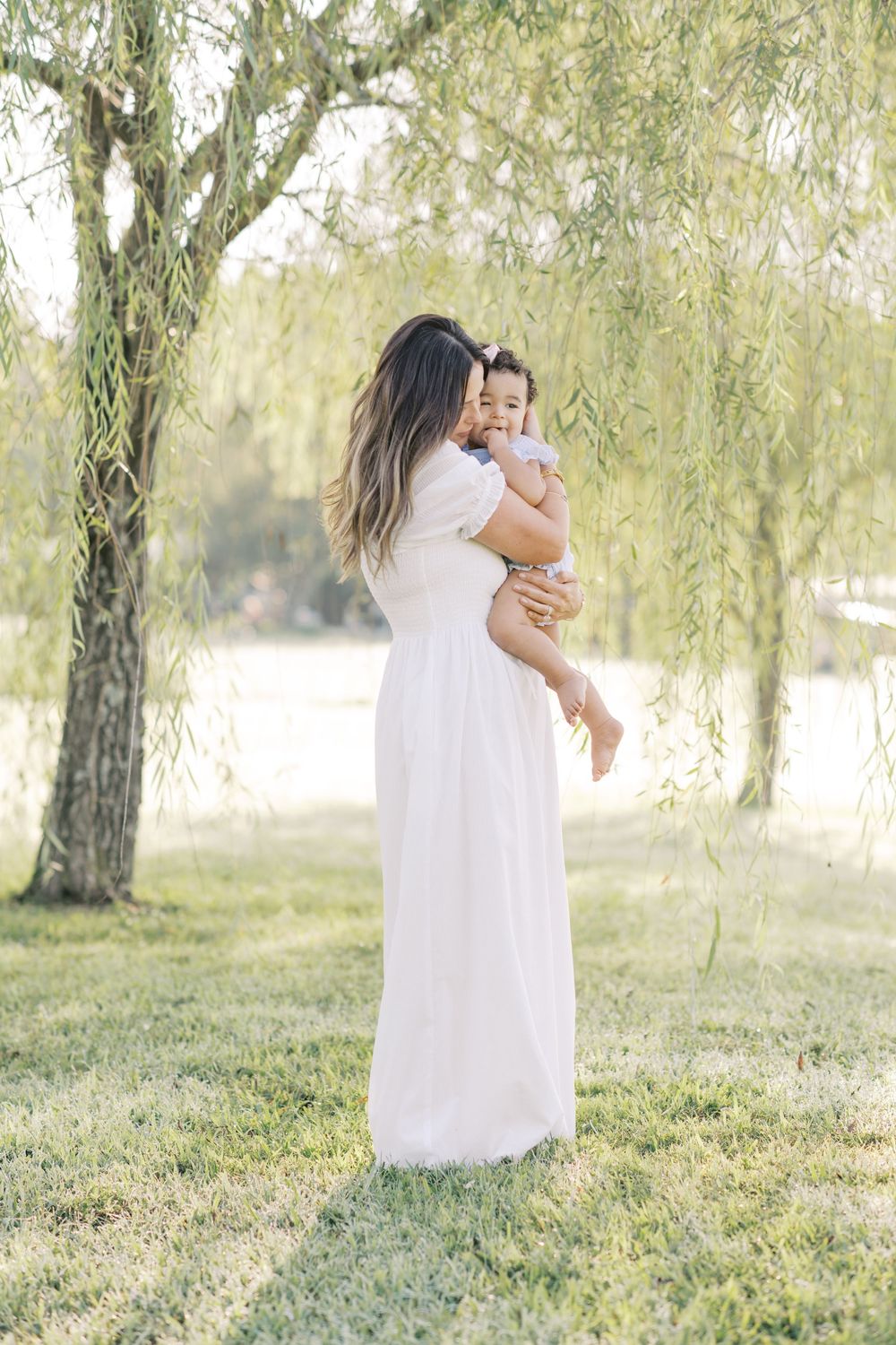 A mother in a white maxi dress holds her baby under a weeping willow tree in a park.