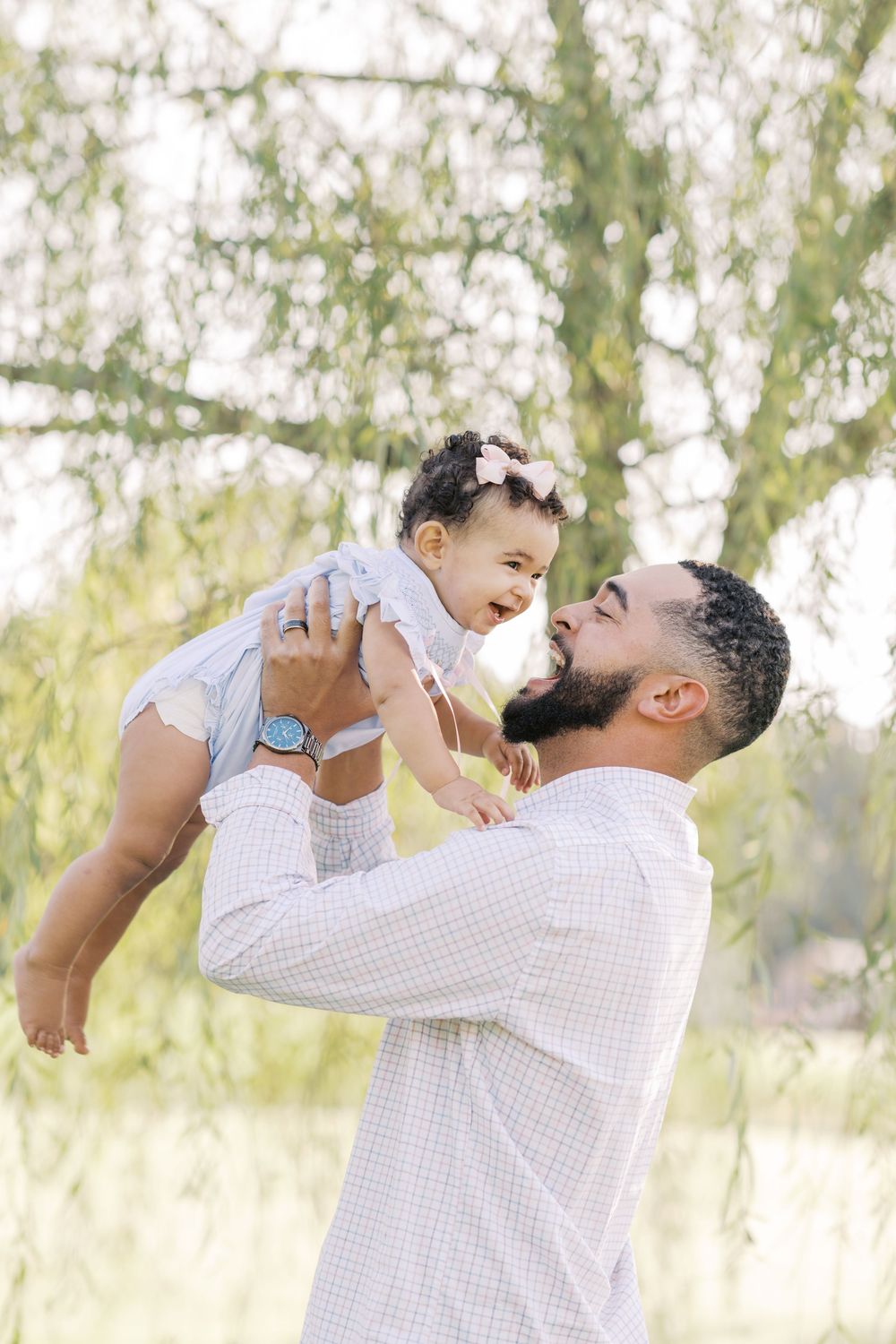 A bearded father lifts his smiling baby girl into the air beneath a leafy tree outdoors.