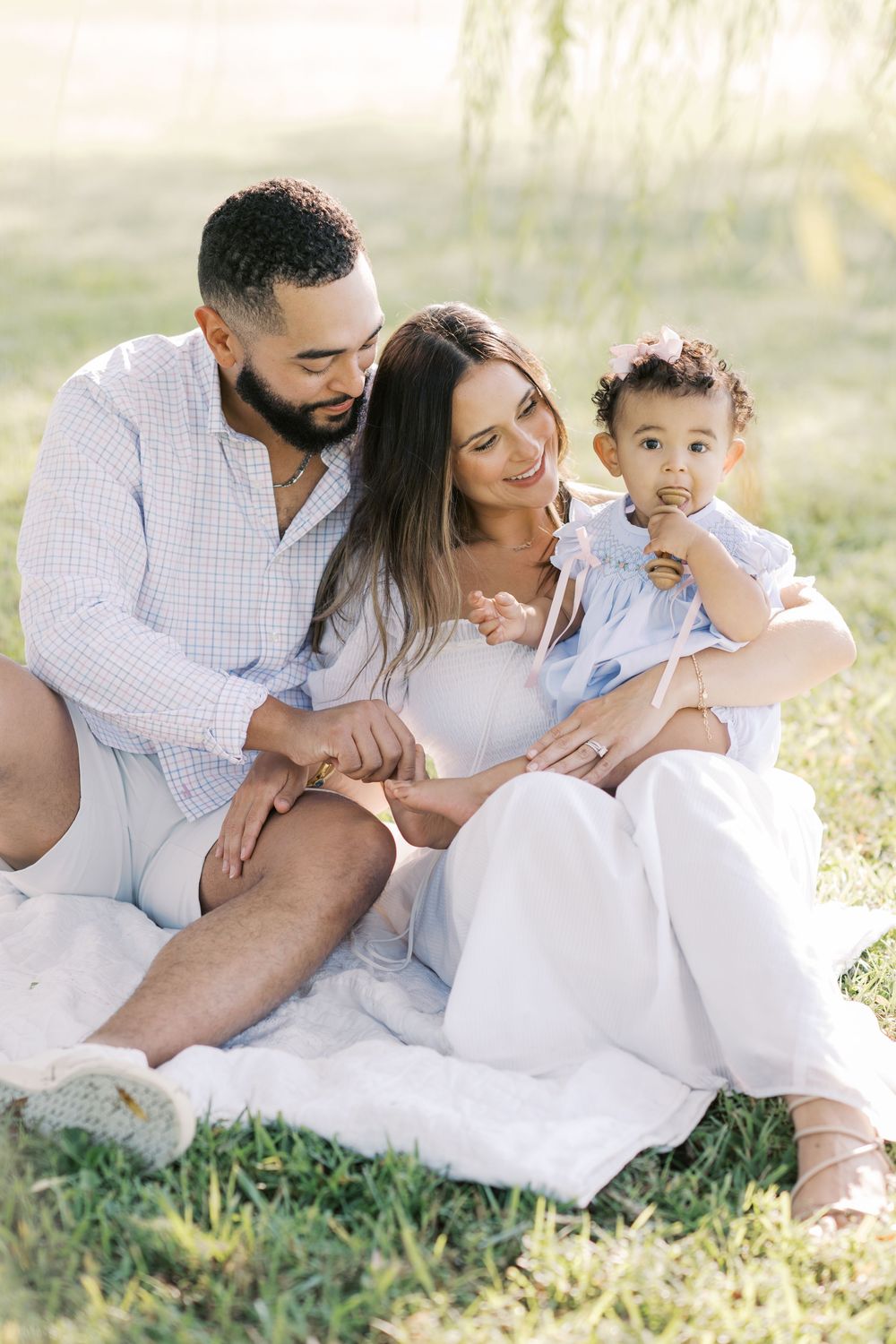 A family sits on a white blanket in the grass as their toddler daughter chews on a toy.