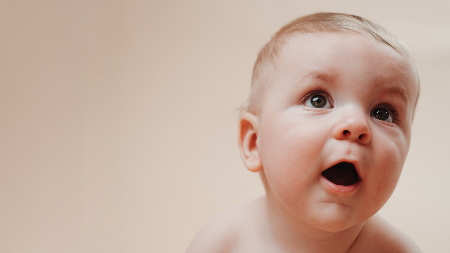 Baby with wide-eyed expression looking up against soft pink background.
