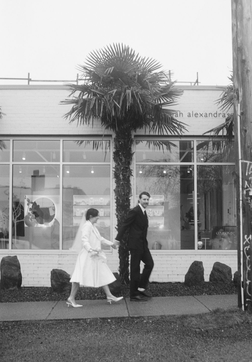 Outdoor black and white scene with palm trees and figures walking along a storefront.
