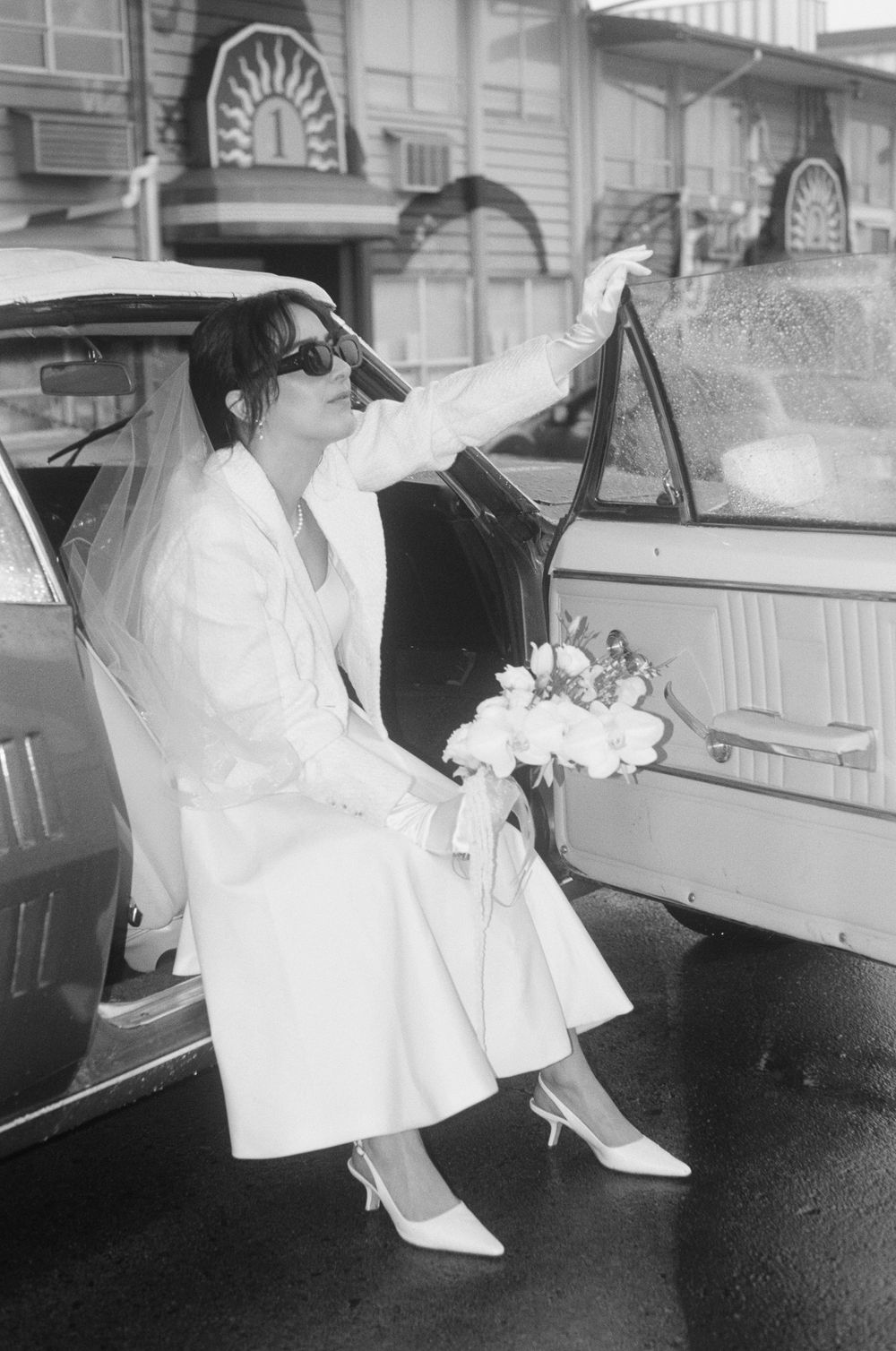 Vintage black and white photo of a bride in white dress entering a classic car.