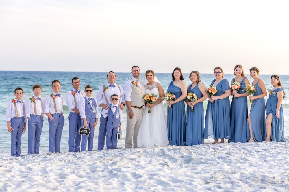 Wedding party wearing blue and white attire poses together on a beautiful white sandy beach by the ocean.