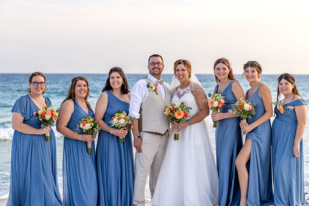 Wedding party in flowing blue dresses and beige suit poses together on beach with ocean waves in background.