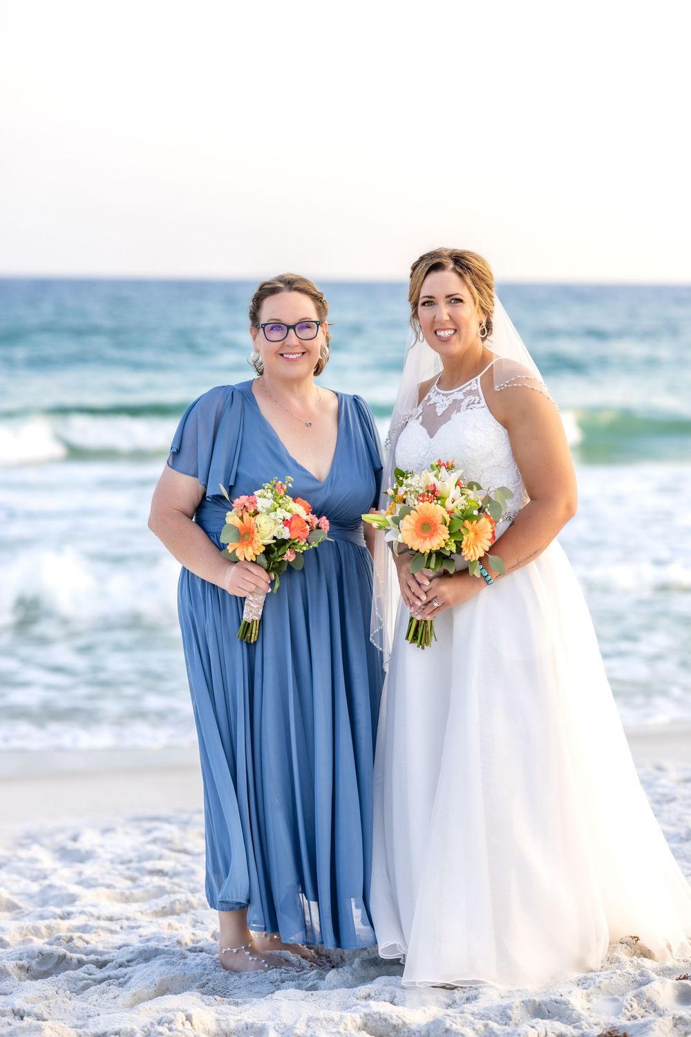 People stand together holding floral bouquets on a sunny beach with ocean waves in the background.