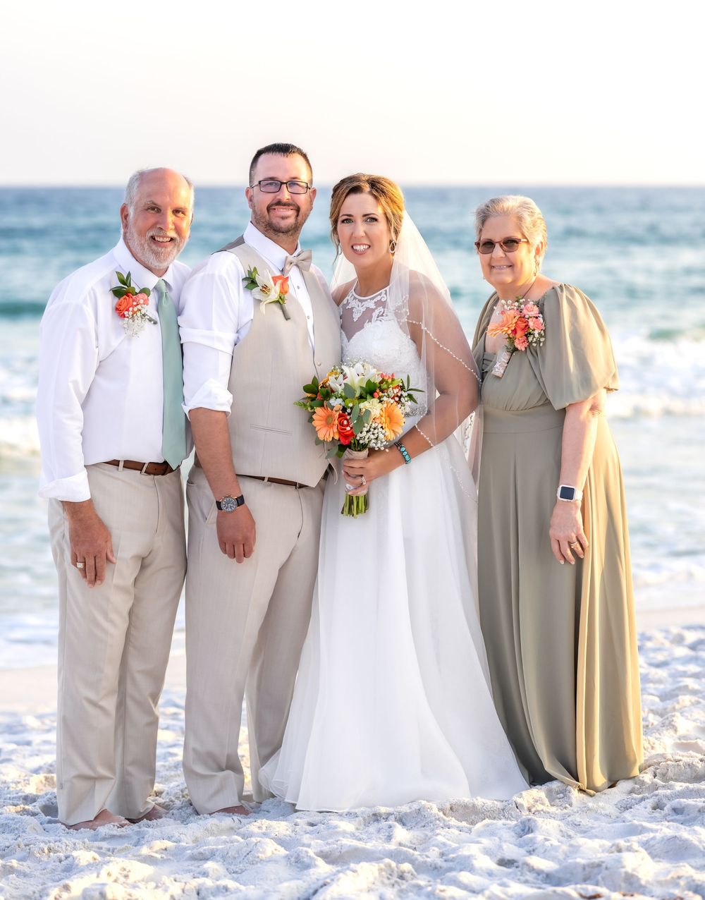 A beach wedding group portrait with four people in light formal attire standing on white sand by the ocean.