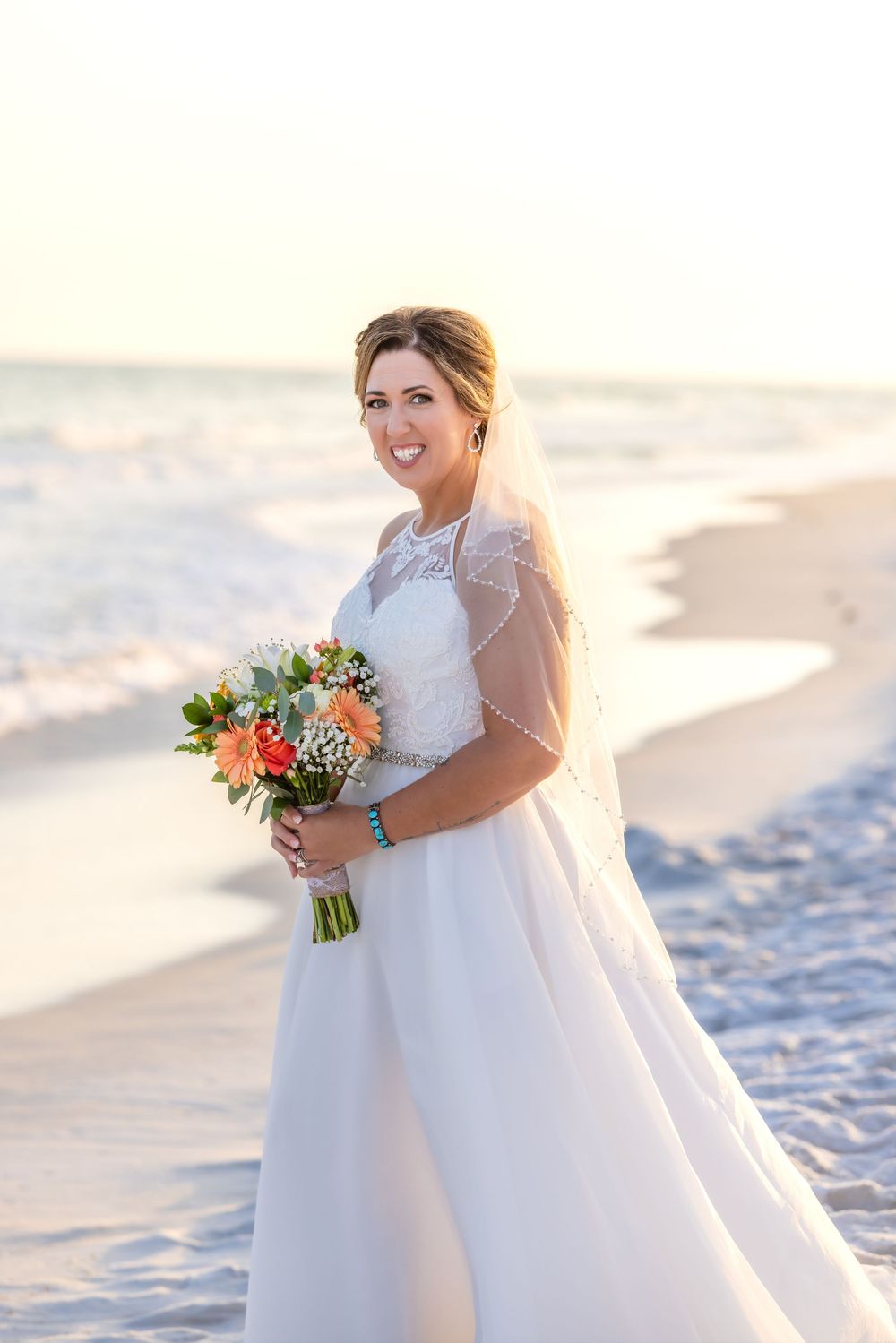 Bride in white wedding dress holding colorful bouquet while standing on sunny beach at sunset.