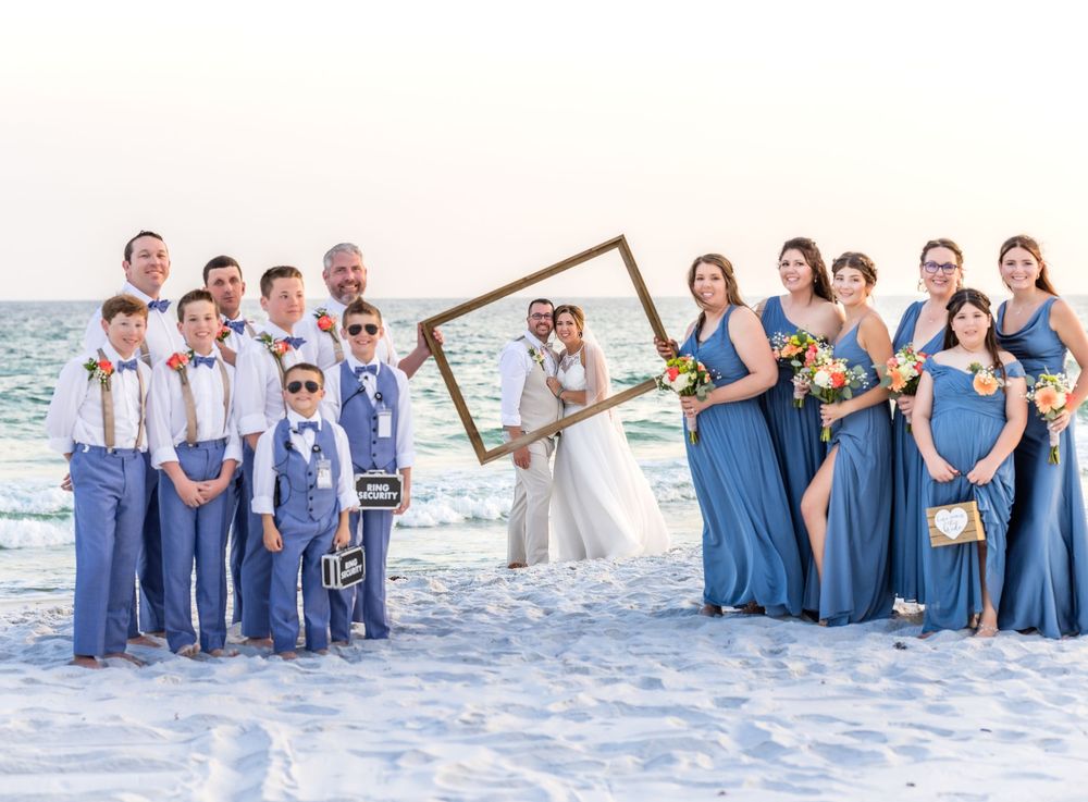 Wedding party in blue and white attire poses with picture frame on sunny beach.