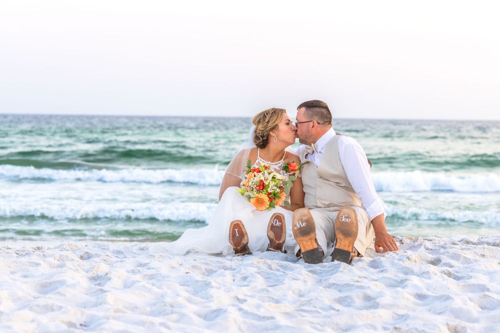 A romantic couple sits together on white sandy beach with turquoise waves rolling in the background.