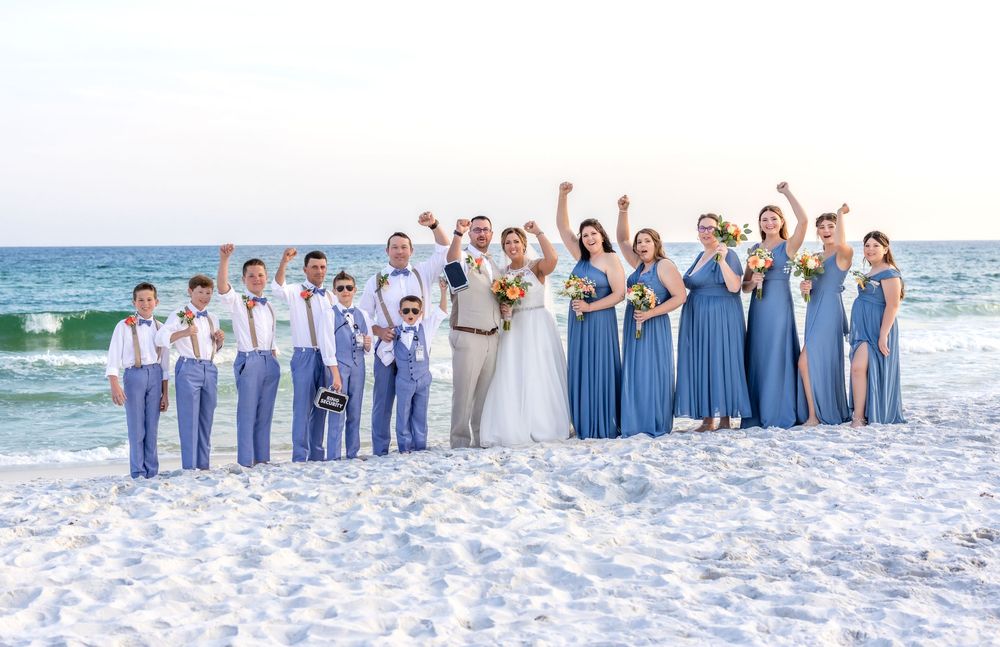 Wedding party in dusty blue dresses and suits poses together on white sandy beach with ocean waves in background.