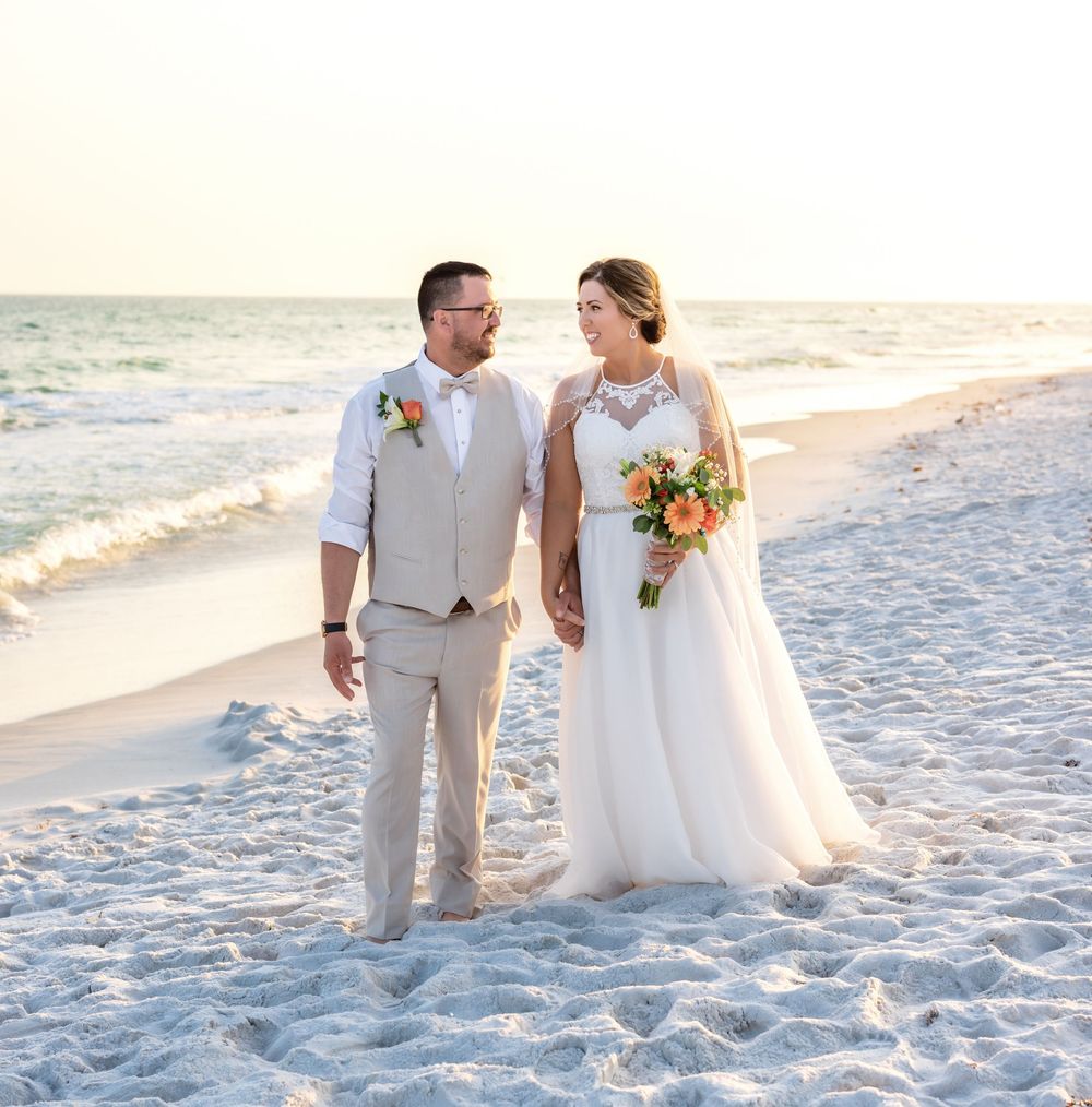 Couple strolls hand in hand on white sandy beach during sunset wedding photoshoot.