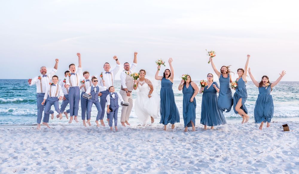 A wedding party celebrates joyfully on a white sandy beach with the ocean waves in the background.