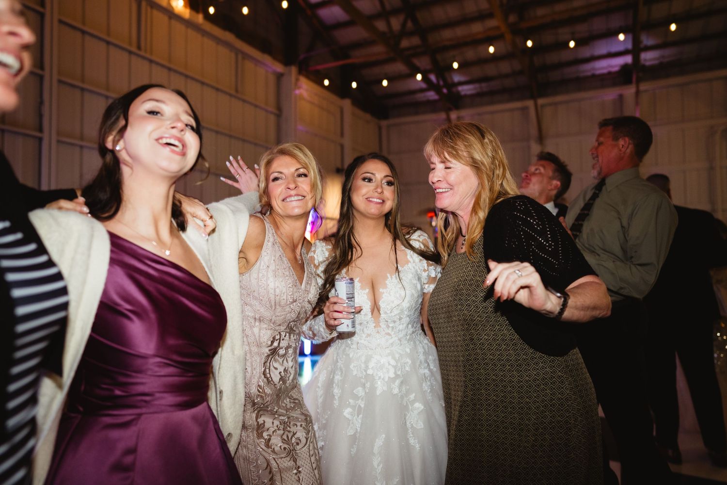 Wedding guests dance and laugh together during a reception celebration in an indoor venue with ambient lighting.