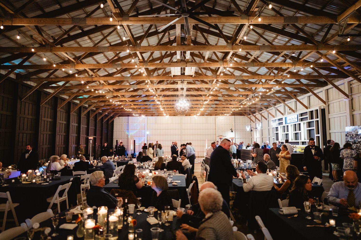 Wedding reception takes place in a rustic barn venue with exposed wooden beams and glowing lights illuminating dining tables.