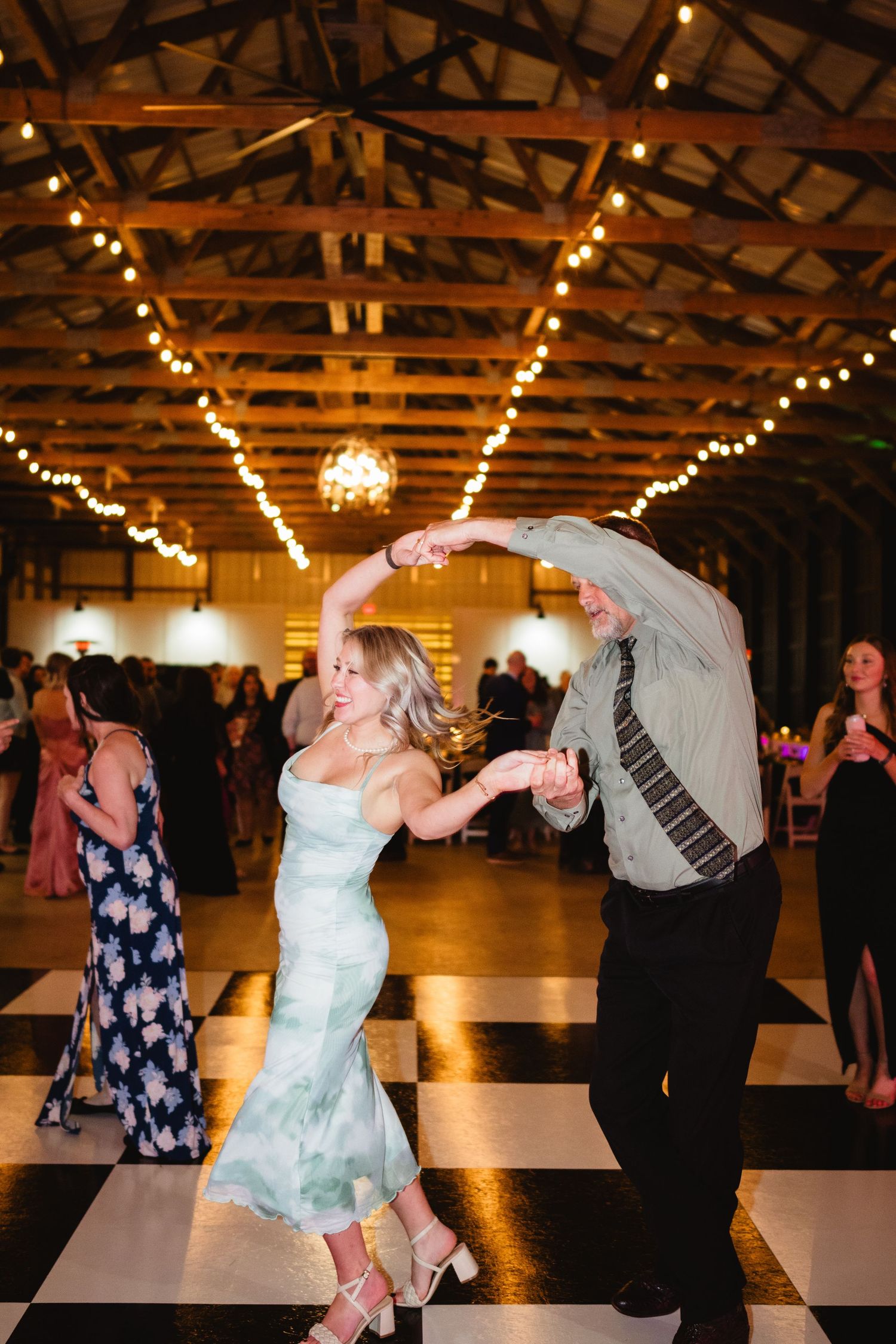 Couple dancing energetically on black and white checkered floor under warm string lights in rustic wooden venue.