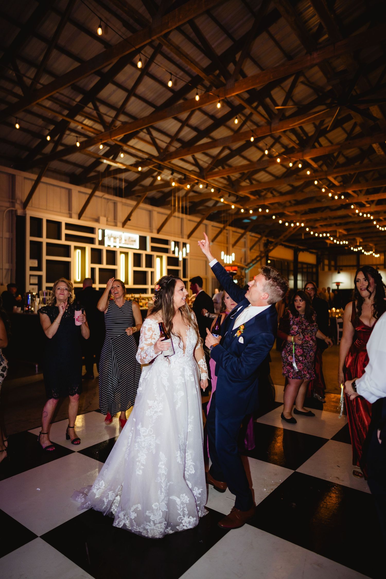 Newlyweds share an energetic dance moment under twinkling lights at their reception venue with black and white flooring.