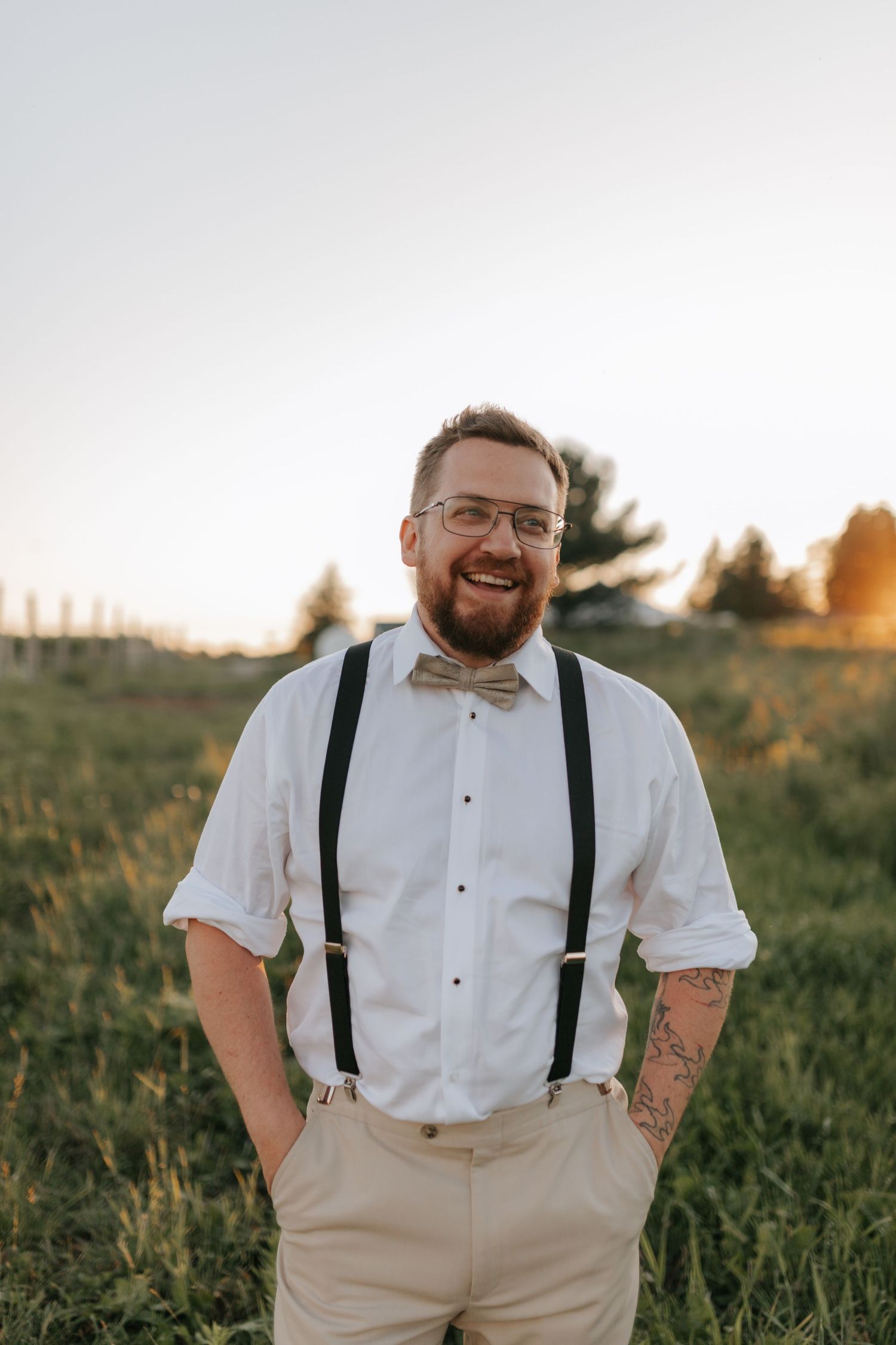Smiling bearded groom in white shirt, suspenders, and bow tie at sunset outdoor wedding.