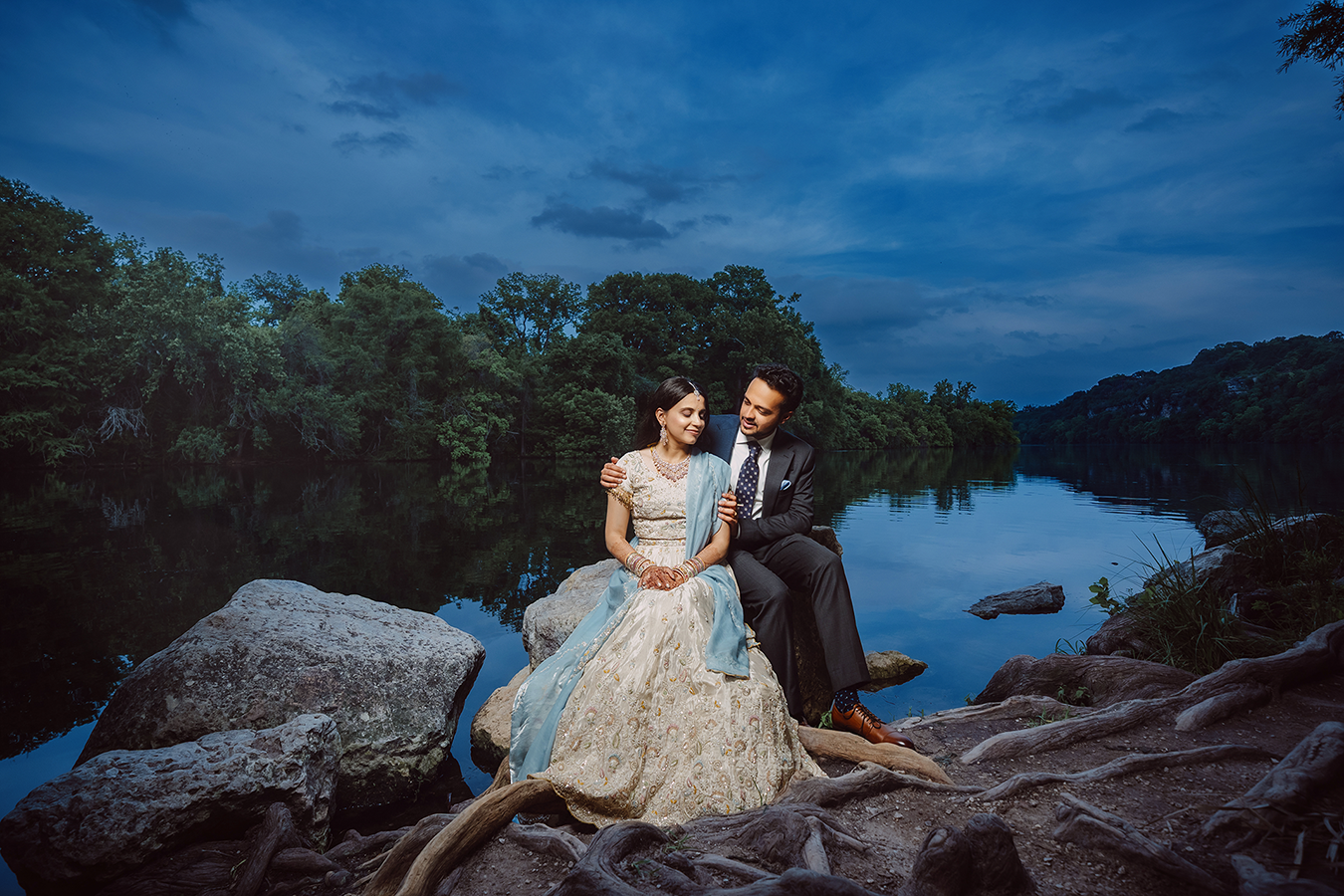 Couple in elegant wedding attire sitting on rocks by a serene river at dusk with lush green trees in background.