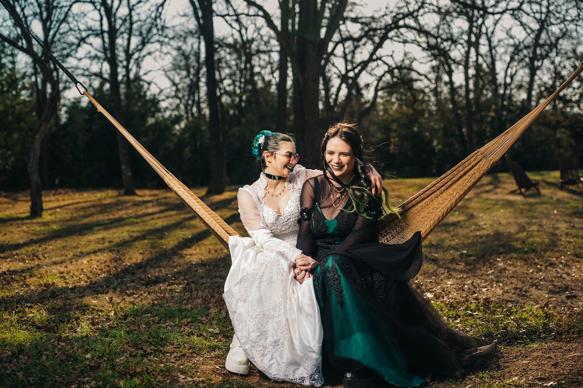 Two brides in white and green gowns sitting together on a hammock outdoors, smiling on their wedding day.