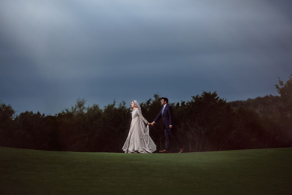 A bride and groom hold hands on a lush green lawn under a dramatic stormy sky at dusk.