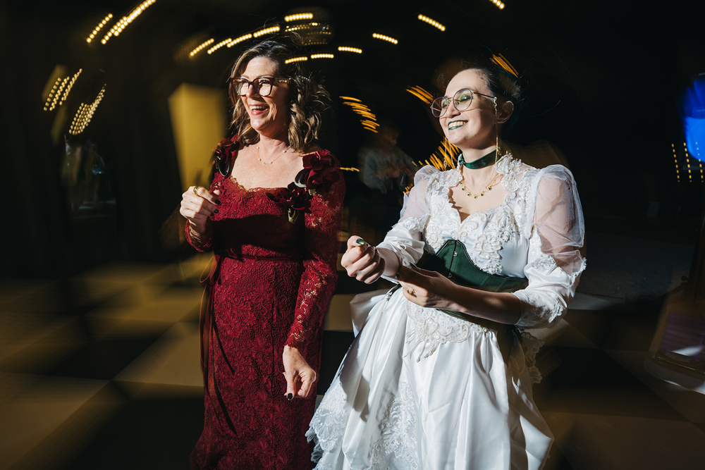 Two women dancing joyfully, one in red lace gown and one in white bridal dress, at an elegant wedding reception.