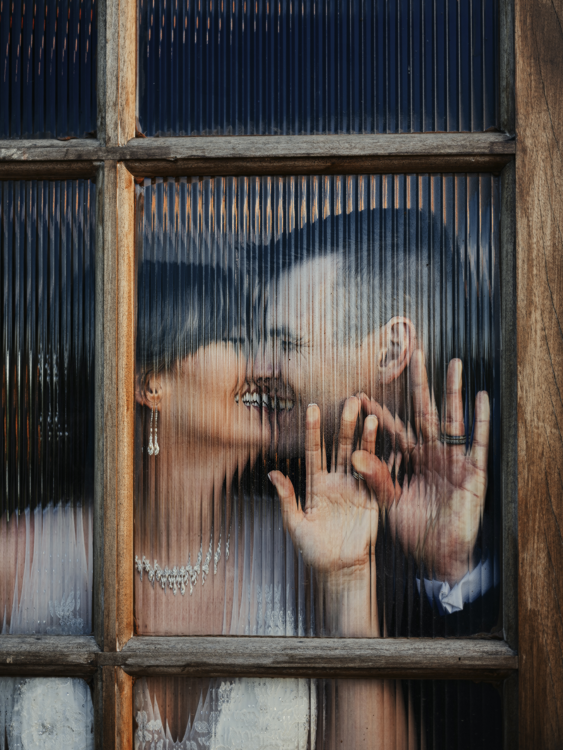 A romantic couple shares an intimate kiss behind a window with vertical blinds creating a soft, dreamy effect.