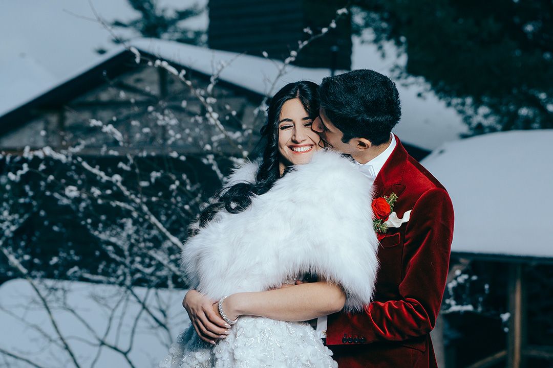 Couple in winter wedding attire embrace in snowy setting, bride in white fur wrap and groom in red velvet suit at Lake Placid