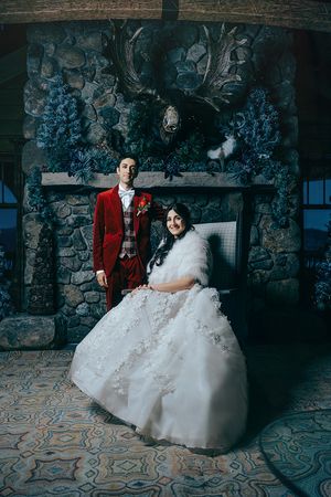 Lake Placid Bride in white gown and groom in red suit pose elegantly before a stone fireplace adorned with blue floral arrangements.