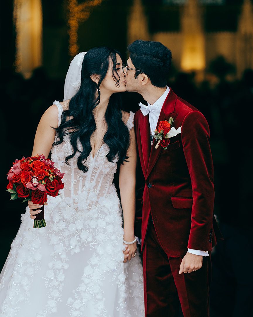Bride and groom share a kiss, she holds red roses bouquet in white floral gown, he wears a red velvet suit.