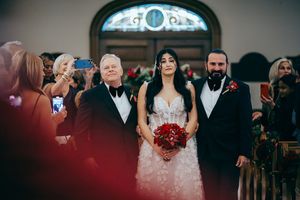 Lake Placid Bride in floral gown holds red bouquet, escorted down church aisle by two men in tuxedos at elegant wedding ceremony.