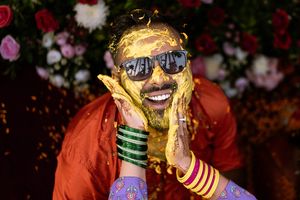 Indian groom covered in yellow turmeric paste smiling at Haldi ceremony, wearing sunglasses and red outfit with green bangles.