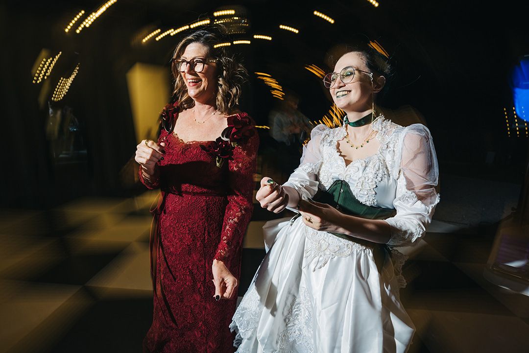 Two women in elegant dresses dance joyfully at a wedding reception, one in red lace and one in white.
