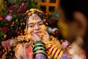 A smiling Indian bride adorned in colorful jewelry and maang tikka during a haldi ceremony with yellow flower petals.