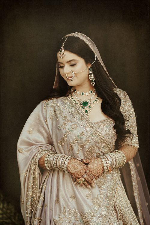 Indian bride in silver lehenga with emerald necklace, maang tikka, bangles, and mehndi poses elegantly.