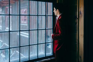 Lake Placid groom in red suit gazing through large grid window, dramatic moody lighting in dark interior setting.